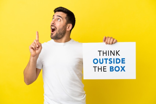 Junger gutaussehender kaukasischer Mann isoliert auf gelbem Hintergrund, der ein Plakat mit dem Text Think Outside The Box hält und denkt
