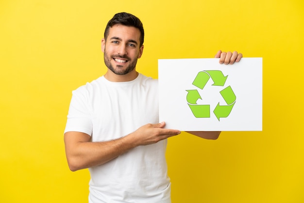 Junger gutaussehender kaukasischer Mann isoliert auf gelbem Hintergrund, der ein Plakat mit Recycling-Symbol mit glücklichem Ausdruck hält