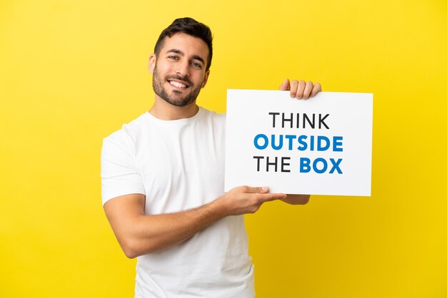 Junger gutaussehender kaukasischer Mann isoliert auf gelbem Hintergrund, der ein Plakat mit Text Think Outside The Box mit glücklichem Ausdruck hält