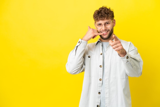 Junger gutaussehender kaukasischer Mann isoliert auf gelbem Hintergrund, der eine Telefongeste macht und nach vorne zeigt