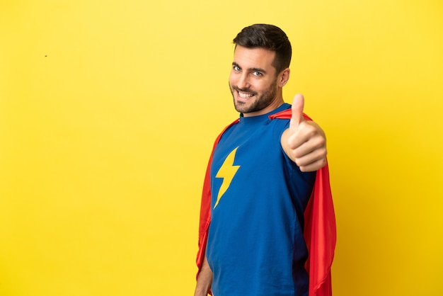 Junger gutaussehender kaukasischer Mann isoliert auf gelbem Hintergrund im Superheldenkostüm mit Daumen nach oben