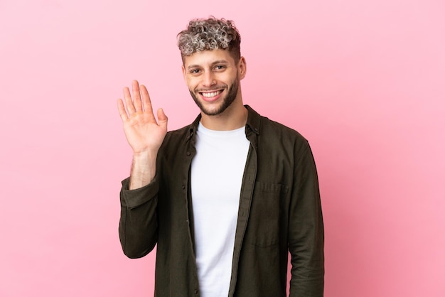 Junger gutaussehender kaukasischer Mann isoliert auf rosa Hintergrund, der mit der Hand mit glücklichem Ausdruck grüßt