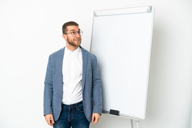 Junger gutaussehender kaukasischer Mann isoliert auf weißem Hintergrund, der eine Präsentation auf dem Whiteboard hält und beim Lächeln aufschaut