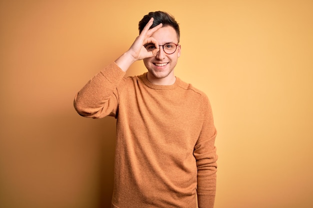 Junger gutaussehender kaukasischer Mann mit Brille und lässigem Winterpullover vor gelbem Hintergrund macht eine OK-Geste mit lächelnder Hand und schaut durch die Finger mit glücklichem Gesicht