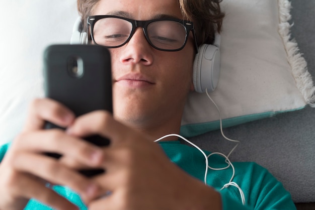 Junger gutaussehender kaukasischer Teenager benutzt ein Mobiltelefon und hört Musik mit Kopfhörern, die sich zu Hause hinlegen - Laden Sie Liederaktivität und modernes vernetztes Lebenskonzept herunter