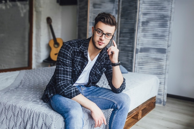 Junger gutaussehender Kerl mit Brille spricht auf einem Handy und posiert in einem Studio im Loft-Stil