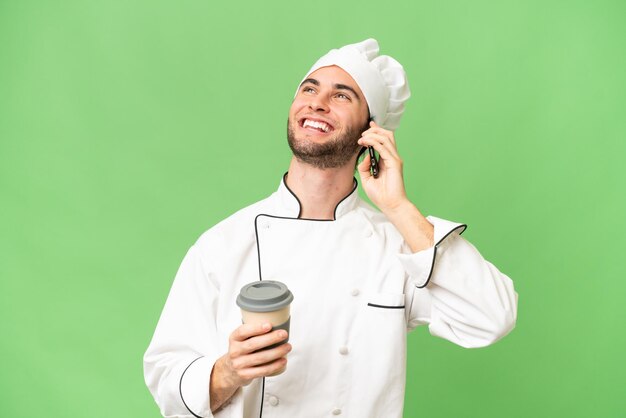 Junger gutaussehender Koch mit isoliertem Hintergrund, der Kaffee zum Mitnehmen und ein Handy in der Hand hält