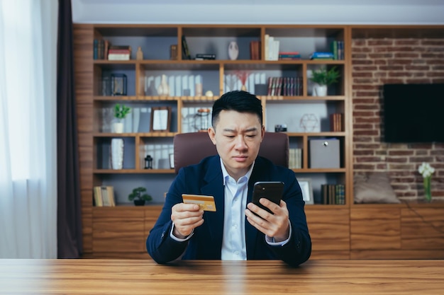 Junger gutaussehender männlicher geschäftsmann bankangestellter direktor asiatisch im anzug im büro am arbeitsplatz hält kreditkarte und nutzt telefon ernst