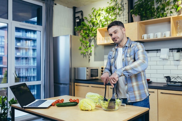 Junger gutaussehender Mann bereitet gesundes und leckeres Essen in der Küche zu Hause zu Essen und Abendessen hackt Gemüse und Obst Er schaut auf die Kamera und lächelt