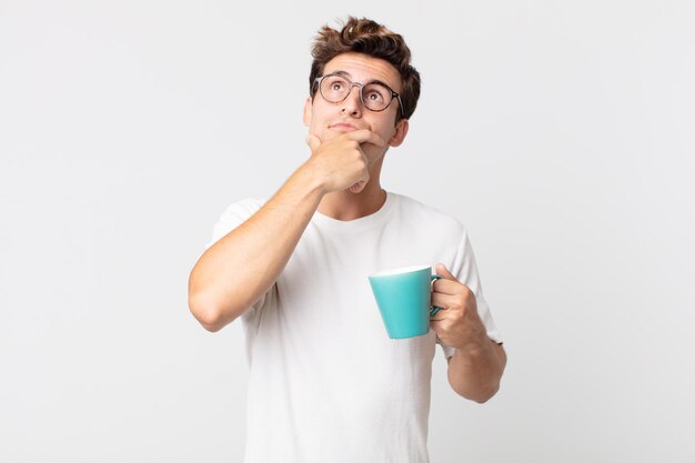 Junger gutaussehender Mann denkt, fühlt sich zweifelnd und verwirrt und hält eine Kaffeetasse