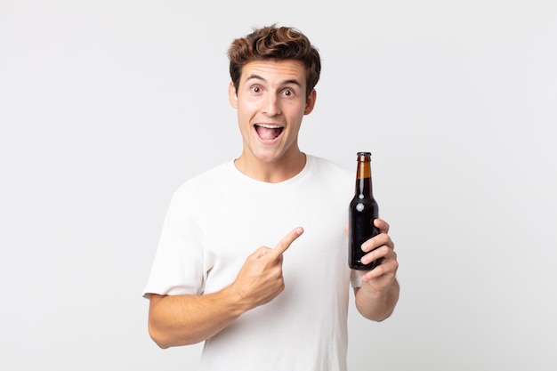 Junger gutaussehender Mann, der aufgeregt und überrascht aussieht, der auf die Seite zeigt und eine Bierflasche hält