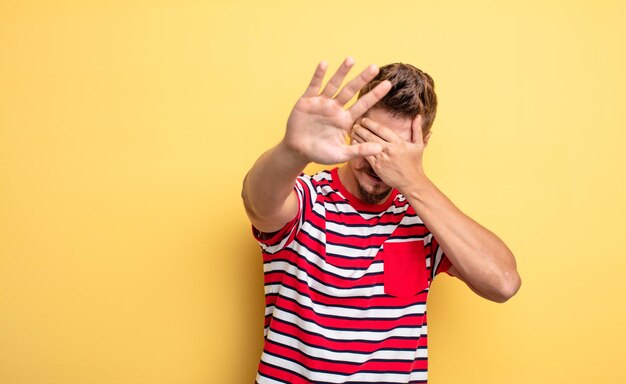 junger gutaussehender mann, der das gesicht mit der hand bedeckt und die andere hand nach vorne legt, um die kamera zu stoppen, fotos oder bilder abzulehnen. seltsames schnurrbartkonzept