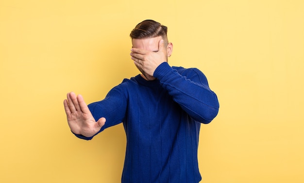 Junger gutaussehender Mann, der das Gesicht mit der Hand bedeckt und die andere Hand nach vorne legt, um die Kamera zu stoppen, Fotos oder Bilder abzulehnen