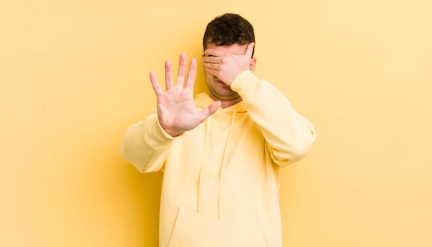 Junger gutaussehender Mann, der das Gesicht mit der Hand bedeckt und die andere Hand nach vorne legt, um zu verhindern, dass die Kamera Fotos oder Bilder ablehnt