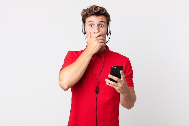 Junger gutaussehender Mann, der den Mund mit den Händen bedeckt, mit einem Schock mit einem Smartphone und einem Headset