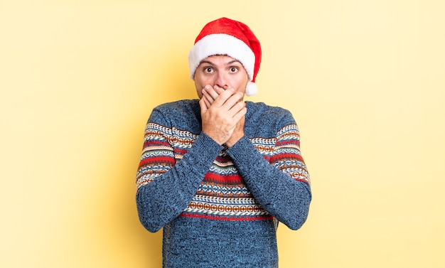 Foto junger gutaussehender mann, der den mund mit den händen mit einem schockierten bedeckt. weihnachtskonzept