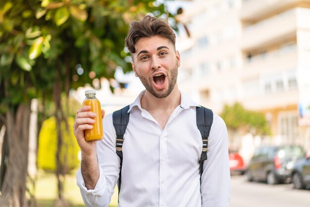 Junger, gutaussehender Mann, der draußen einen Orangensaft in der Hand hält, mit überraschtem und schockiertem Gesichtsausdruck