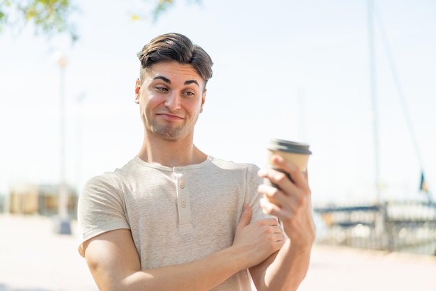 Junger gutaussehender Mann, der draußen mit traurigem Ausdruck einen Kaffee zum Mitnehmen hält