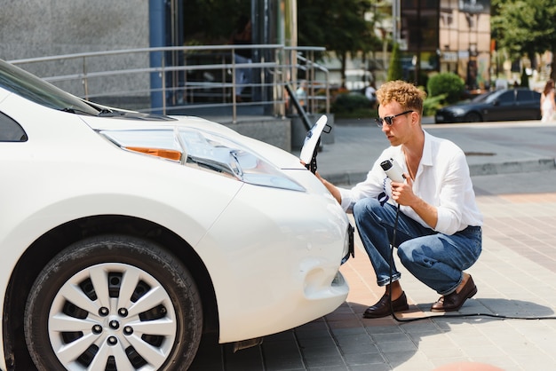 Junger gutaussehender Mann, der ein Ladekabel an einer elektrischen Ladestation hält, der in der Nähe seines neuen Autos steht und zufrieden aussieht.