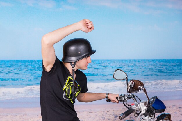 Junger gutaussehender Mann, der ein Motorrad am Strand mit einem Arm reitet, der auf einem blauen ruhigen Ozeanhintergrund angehoben wird.