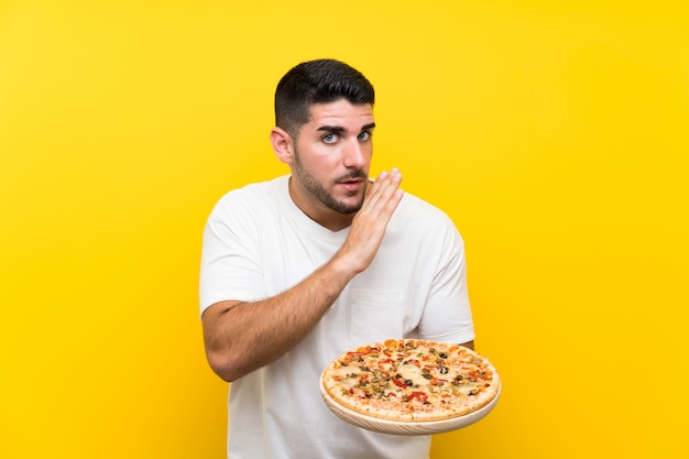 Junger gutaussehender mann, der eine pizza über der lokalisierten gelben wand flüstert etwas hält