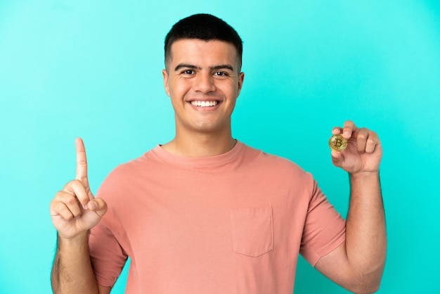 Junger gutaussehender Mann, der einen Bitcoin über isoliertem blauem Hintergrund hält und einen Finger im Zeichen des Besten zeigt und hebt