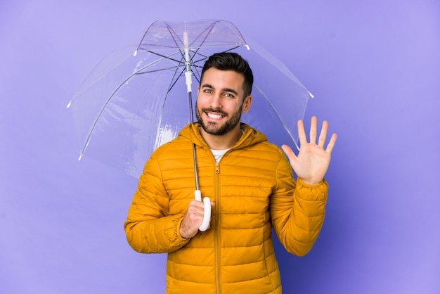 Junger gutaussehender Mann, der einen Regenschirm isoliert hält, der lächelnd fröhlich zeigt Nummer fünf mit den Fingern.