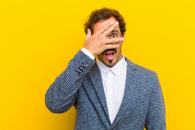 Junger gutaussehender Mann, der entsetzt, erschrocken oder erschrocken schaut, Gesicht mit der Hand bedeckt und zwischen Finger gegen Orange späht