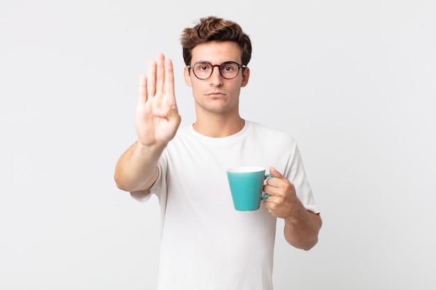 Junger gutaussehender Mann, der ernst aussieht und eine offene Handfläche zeigt, die Stopp-Geste macht und eine Kaffeetasse hält