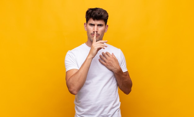 Junger gutaussehender Mann, der ernst aussieht und mit dem Finger auf die Lippen drückt, die Stille oder Ruhe fordern und ein Geheimnis bewahren