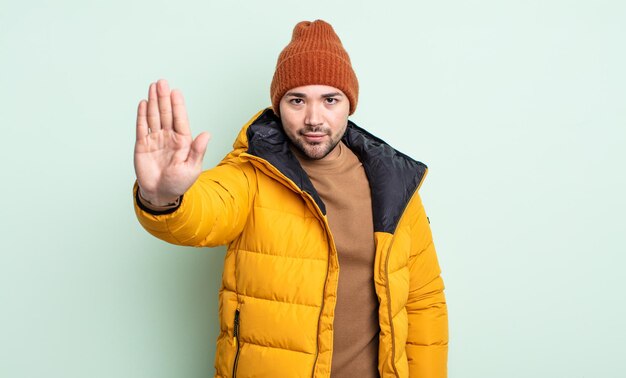 Junger gutaussehender Mann, der ernst aussieht und offene Handfläche zeigt, die Stopp-Geste macht. Konzept für kaltes Wetter