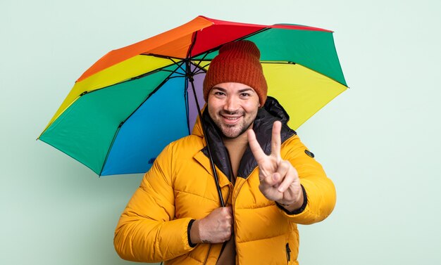 Junger gutaussehender Mann, der freundlich lächelt und aussieht und Nummer zwei zeigt. Regen- und Regenschirmkonzept