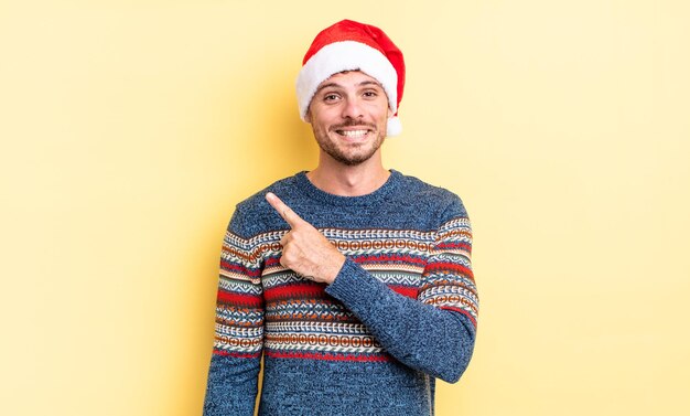 Junger gutaussehender Mann, der fröhlich lächelt, sich glücklich fühlt und auf die Seite zeigt. Weihnachtskonzept