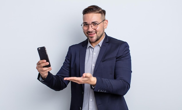 Junger gutaussehender Mann, der fröhlich lächelt, sich glücklich fühlt und ein Konzeptgeschäft und ein Smartphone-Konzept zeigt