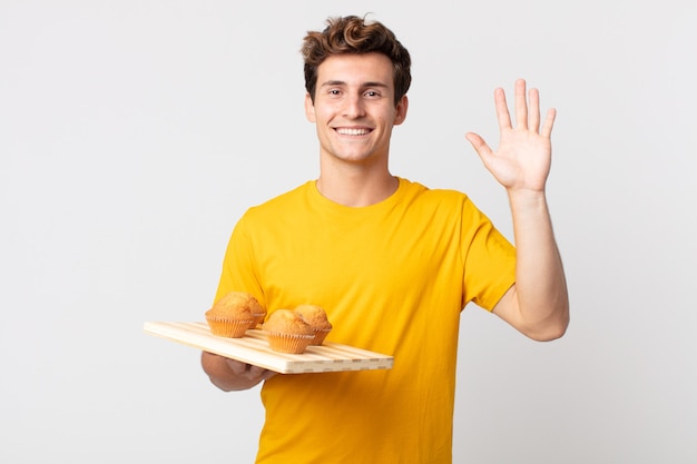 Junger gutaussehender Mann, der glücklich lächelt, mit der Hand winkt, Sie begrüßt und begrüßt, der ein Muffins-Tablett hält?