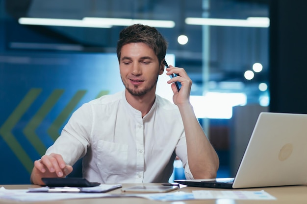 Junger gutaussehender Mann, der im Büro am Schreibtisch arbeitet und mit Kunden bei der Bank mit Geschäftspartnern über ein Mobiltelefon spricht