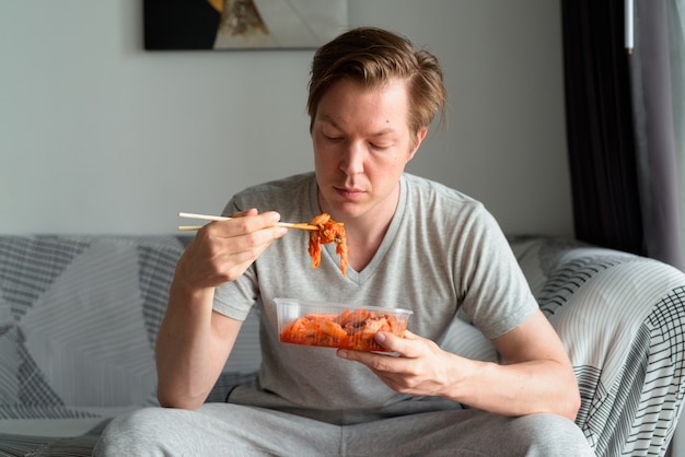 Junger gutaussehender Mann, der Kimchi isst, während er auf dem Sofa zu Hause sitzt