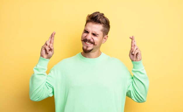 Junger gutaussehender Mann, der lächelt und ängstlich beide Finger drückt, sich besorgt fühlt und auf ein seltsames Schnurrbart-Konzept wünscht oder hofft