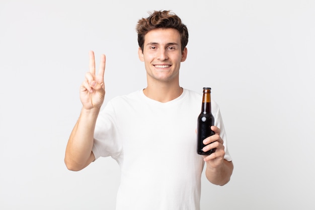 Junger gutaussehender Mann, der lächelt und freundlich aussieht, Nummer zwei zeigt und eine Bierflasche hält?