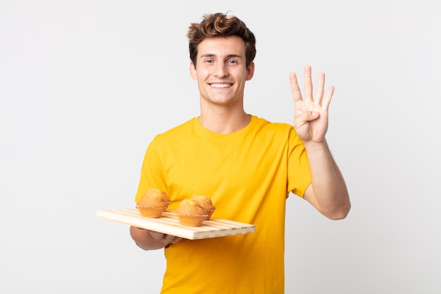 Junger gutaussehender Mann, der lächelt und freundlich aussieht und die Nummer vier zeigt, die ein Muffins-Tablett hält?