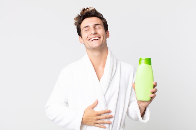 Junger gutaussehender Mann, der laut über einen lustigen Witz mit Bademantel lacht und eine Shampooflasche hält