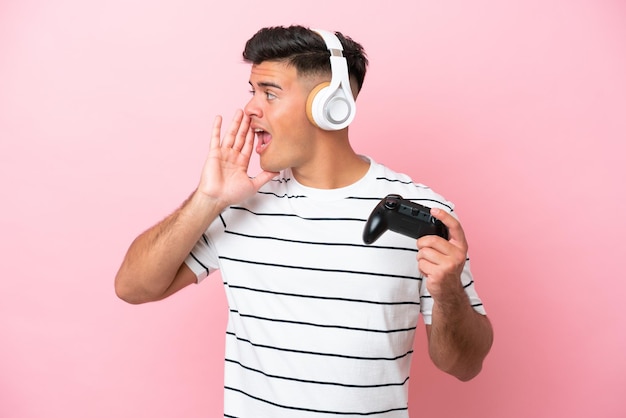 Junger gutaussehender Mann, der mit einem Videospiel-Controller spielt, der isoliert auf rosafarbenem Hintergrund schreit, mit weit geöffnetem Mund zur Seite