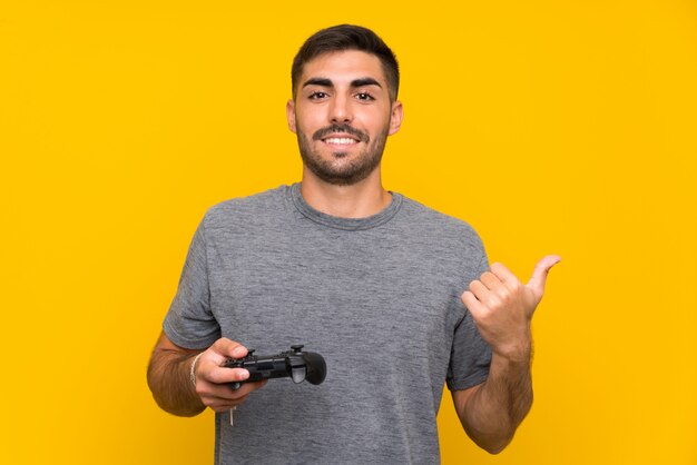 Junger gutaussehender Mann, der mit einem Videospielcontroller über lokalisierter gelber Wand zeigt auf die Seite spielt, um ein Produkt darzustellen