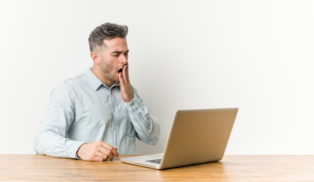 Junger gutaussehender Mann, der mit seinem Laptop gähnt arbeitet, eine müde Geste zeigend, die Mund mit der Hand bedeckt.