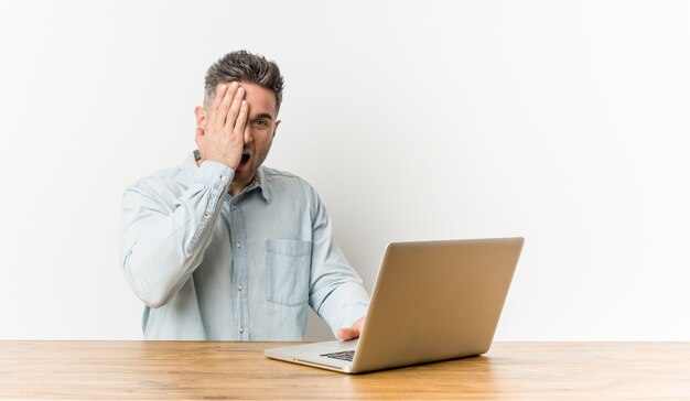 Junger gutaussehender Mann, der mit seinem Laptop hat den Spaß bedeckt die Hälfte des Gesichtes mit Palme arbeitet.