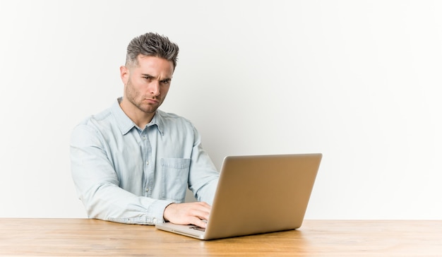 Junger gutaussehender Mann, der mit seinem Laptop missbilligend das Stirnrunzeln arbeitet, hält Arme verschränkt.