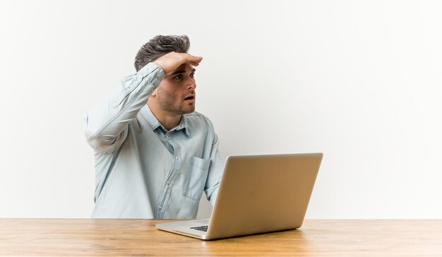 Junger gutaussehender Mann, der mit seinem Laptop schaut weit weg arbeitet, Hand auf Stirn halten.