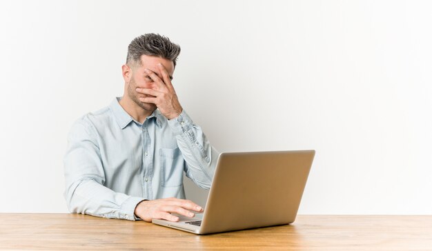 Junger gutaussehender Mann, der mit seinem Laptopblinken durch Finger, verlegenes Bedeckungsgesicht arbeitet.