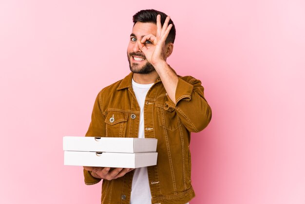 Junger gutaussehender Mann, der Pizzas lokalisiert aufgeregt hält und ok Geste auf Auge hält.