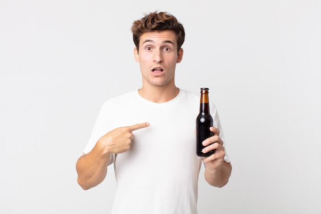 Junger gutaussehender Mann, der schockiert und überrascht mit weit geöffnetem Mund aussieht, auf sich selbst zeigt und eine Bierflasche hält